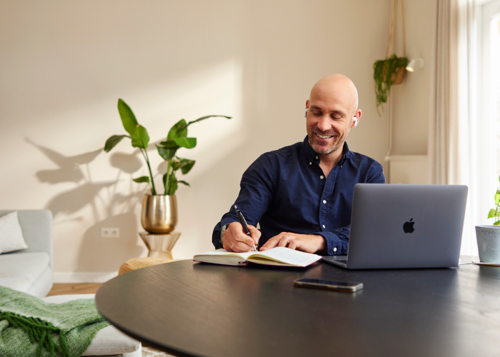 Een man zit aan tafel te schrijven achter zijn laptop