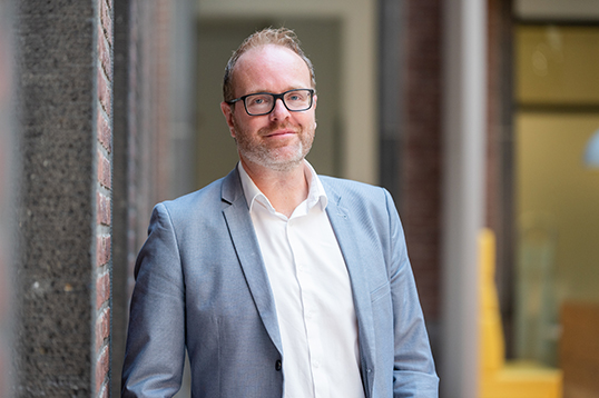 Man met licht blauw pak en zwart bril glimlacht.